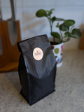 Black coffee bag with a label on a countertop with a plant in the background